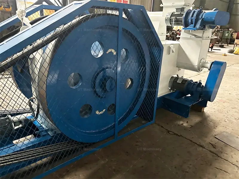 Grande volante de inércia azul e sistema de correia de transmissão com uma malha de proteção de segurança em uma máquina briquetadeira de biomassa industrial.