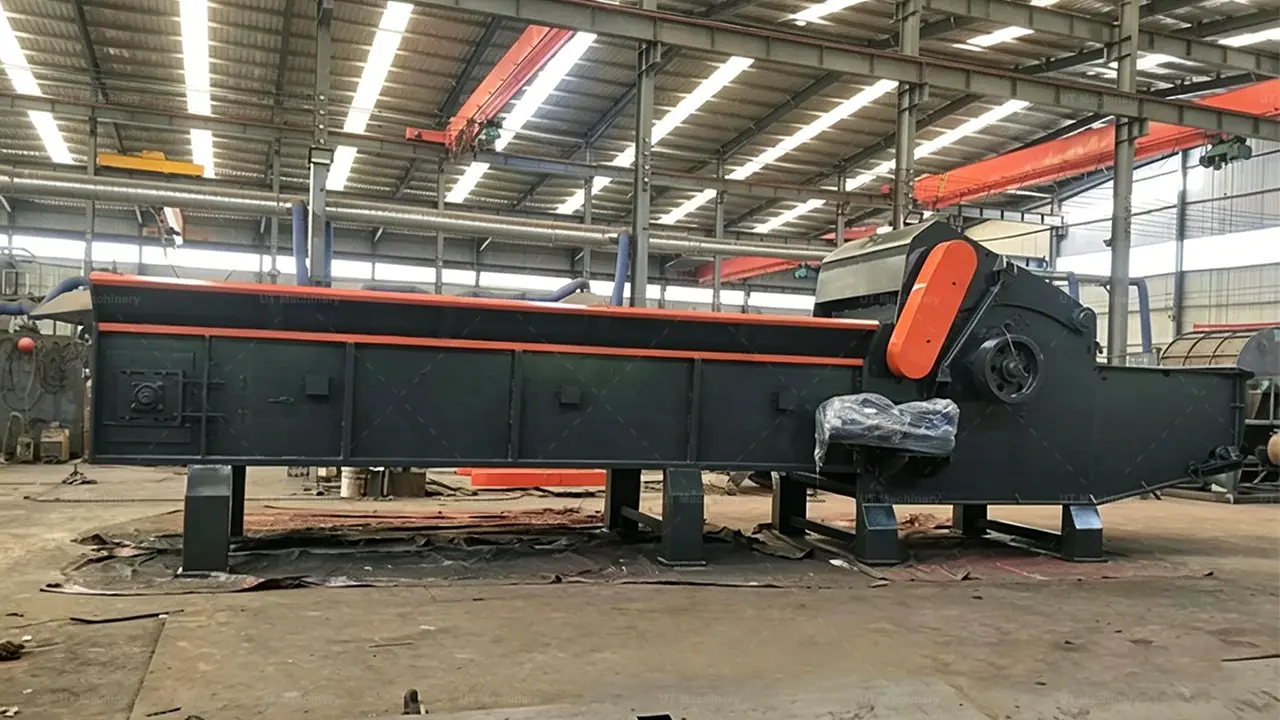 Foto de perfil lateral a longa distância e ao nível dos olhos de um enorme picador de madeira industrial cinza-escuro dentro de uma oficina bem iluminada. A máquina apresenta uma calha de alimentação baixa e muito longa à esquerda com um sistema de alimentação de lona de corrente integrado, levando à grande unidade de processamento à direita.